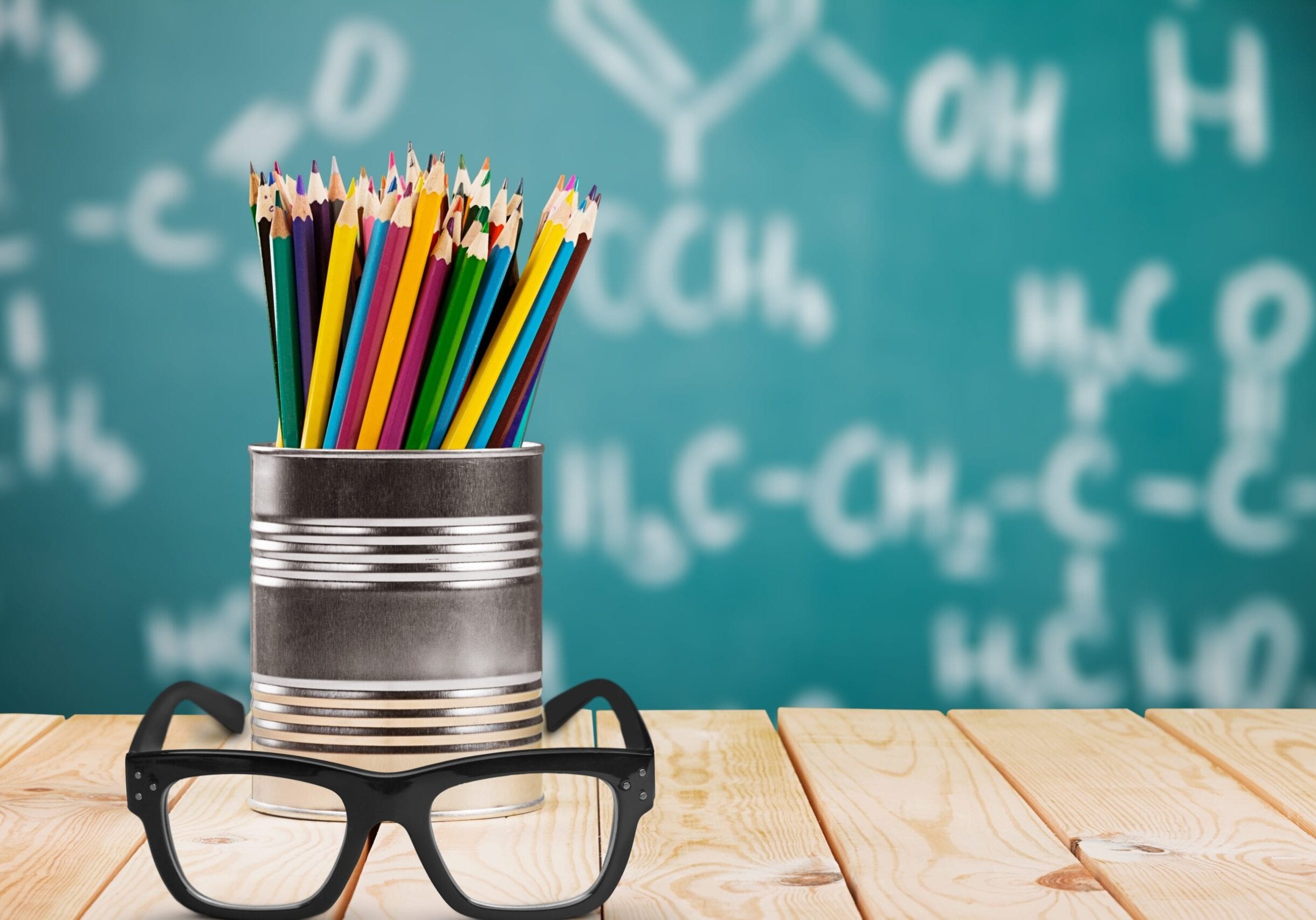Classroom image: Pot of pencils and pair of glasses on desk in foreground with blackboard with writing on it blurred in background